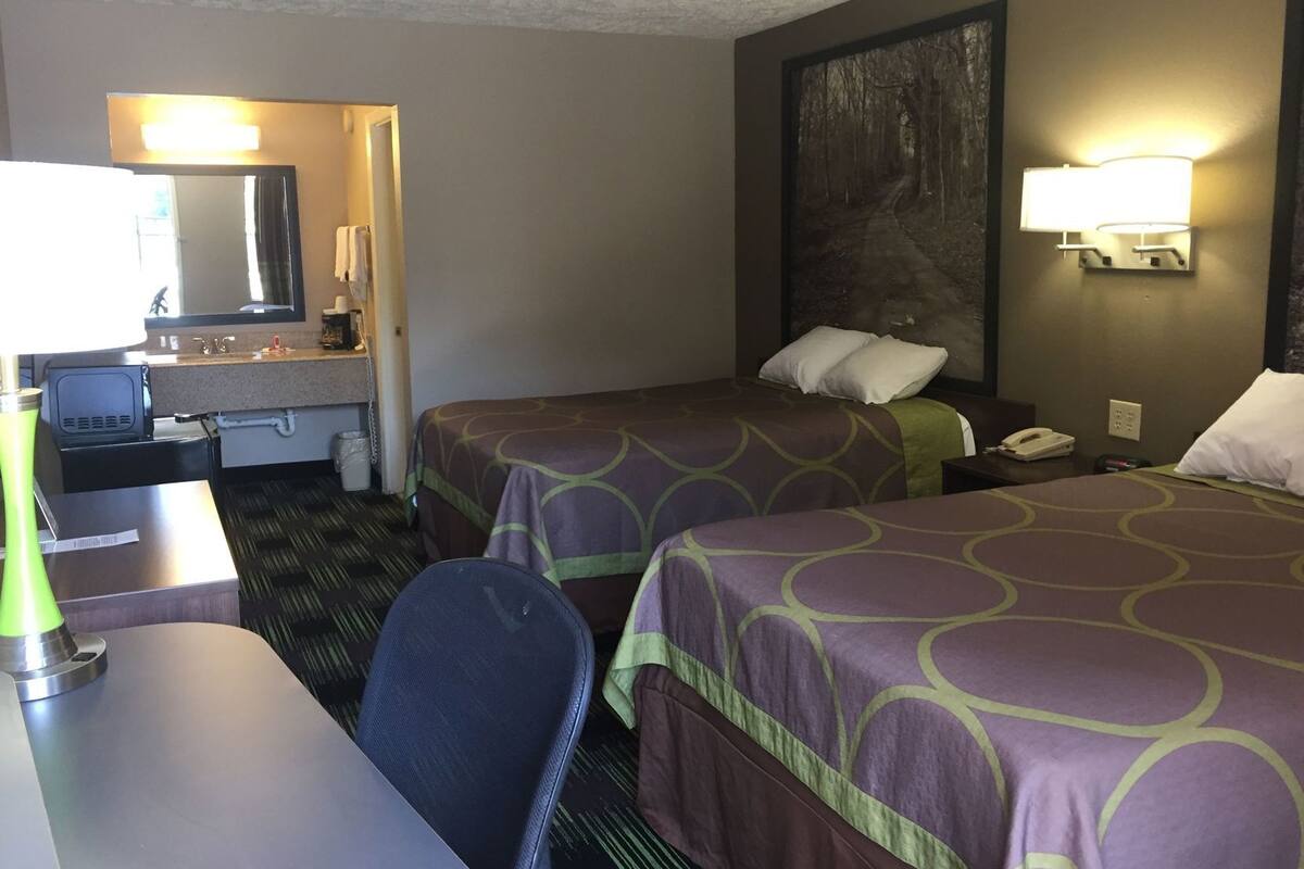 A hotel room with two beds, a desk, a chair, a desk lamp, a telephone, and a wall-mounted light fixture.