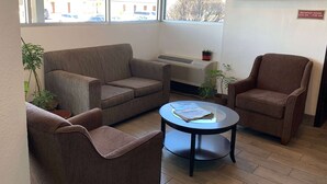 Una habitación con un sofá marrón, un sillón marrón y una mesa redonda con una revista. Se ve una ventana con vista a la calle y un letrero que indica los horarios del desayuno.