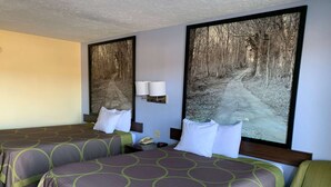 Habitación de hotel con dos camas, una mesita de noche, una lámpara y un cuadro grande con temática de bosque en la pared.