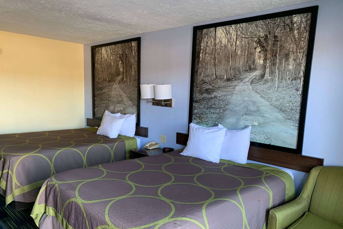 A hotel room with two beds, a nightstand, a lamp, and a large forest-themed painting on the wall.