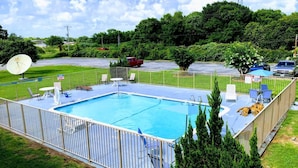A rectangular swimming pool with a white fence, surrounded by a grassy area with a few trees and a satellite dish. - Motel 6 Clute, TX (Clute)