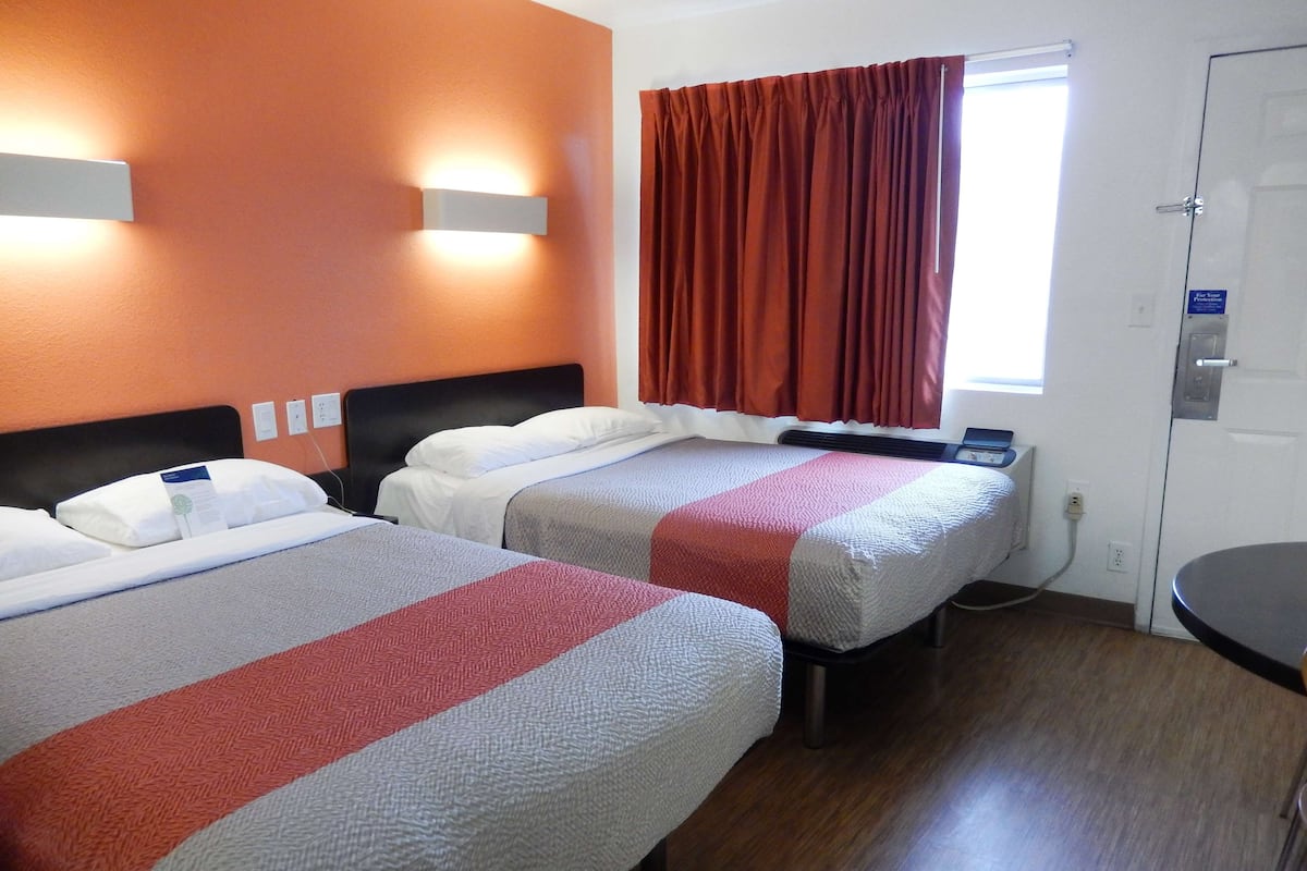 A hotel room with two beds, a window with curtains, a bedside table, and a door.