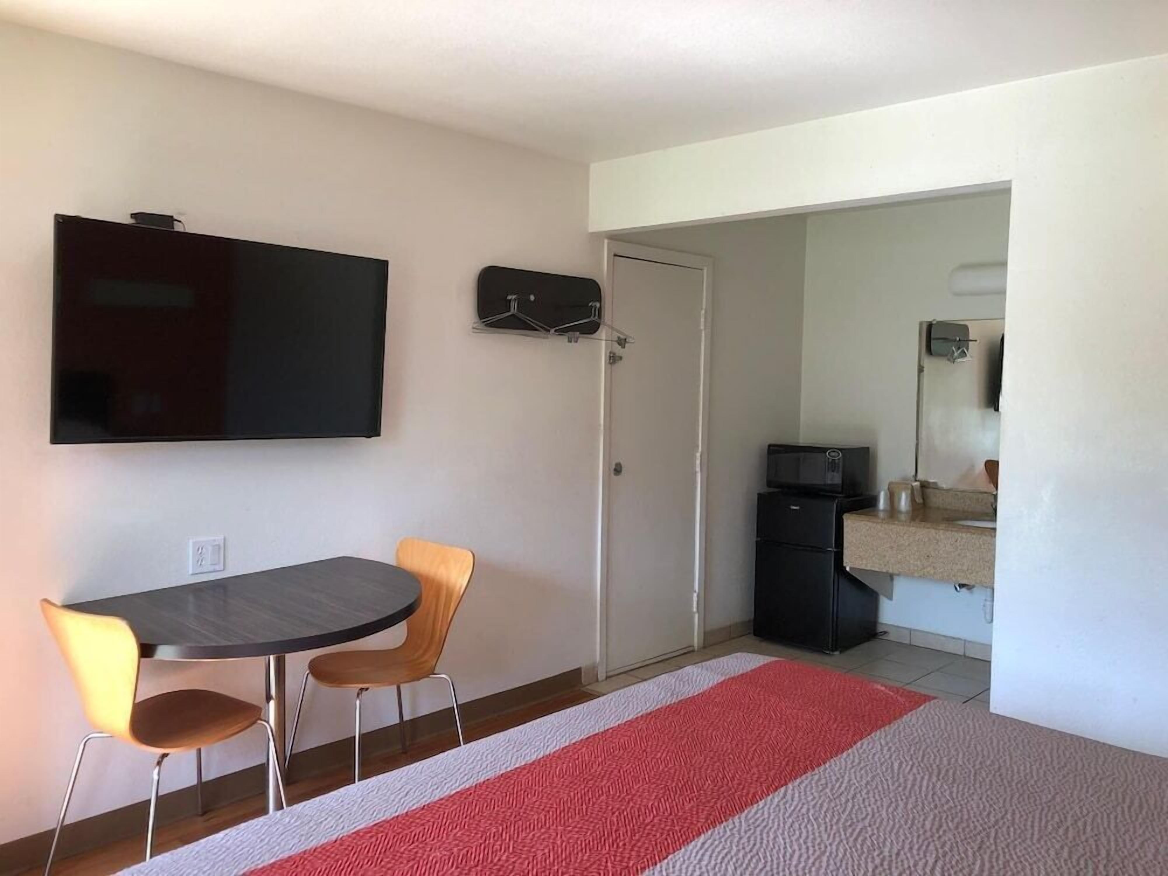 A small room with a dining table, two chairs, a flat-screen TV mounted on the wall, a microwave, and a refrigerator in the corner.