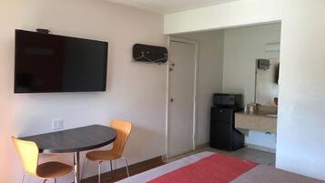 Una habitación pequeña con una mesa de comedor, dos sillas, un televisor de pantalla plana en la pared, un microondas y un refrigerador en la esquina.