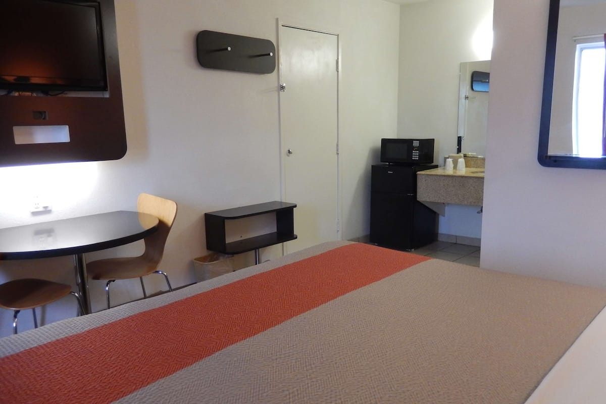 A hotel room with a bed, a desk with two chairs, a small refrigerator, a television mounted on the wall, and a window.
