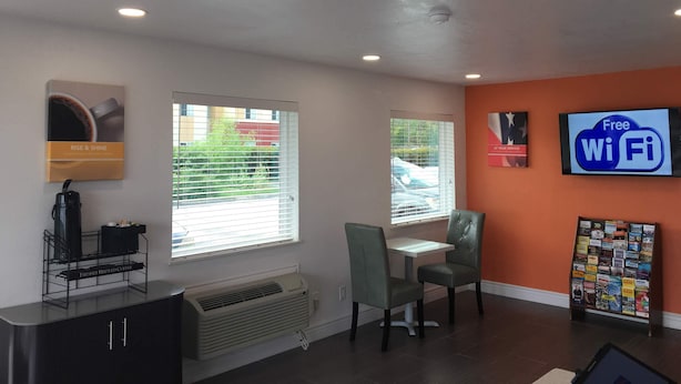 A modern interior with a coffee station, two windows with blinds, a table and chairs, a Wi-Fi sign, and a magazine rack. - Motel 6 Clute, TX (Clute)