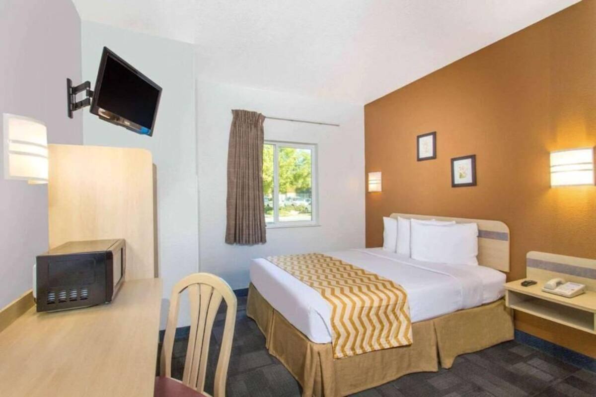 Quarto de hotel com cama, escrivaninha com computador, televisão na parede e janela com cortinas.