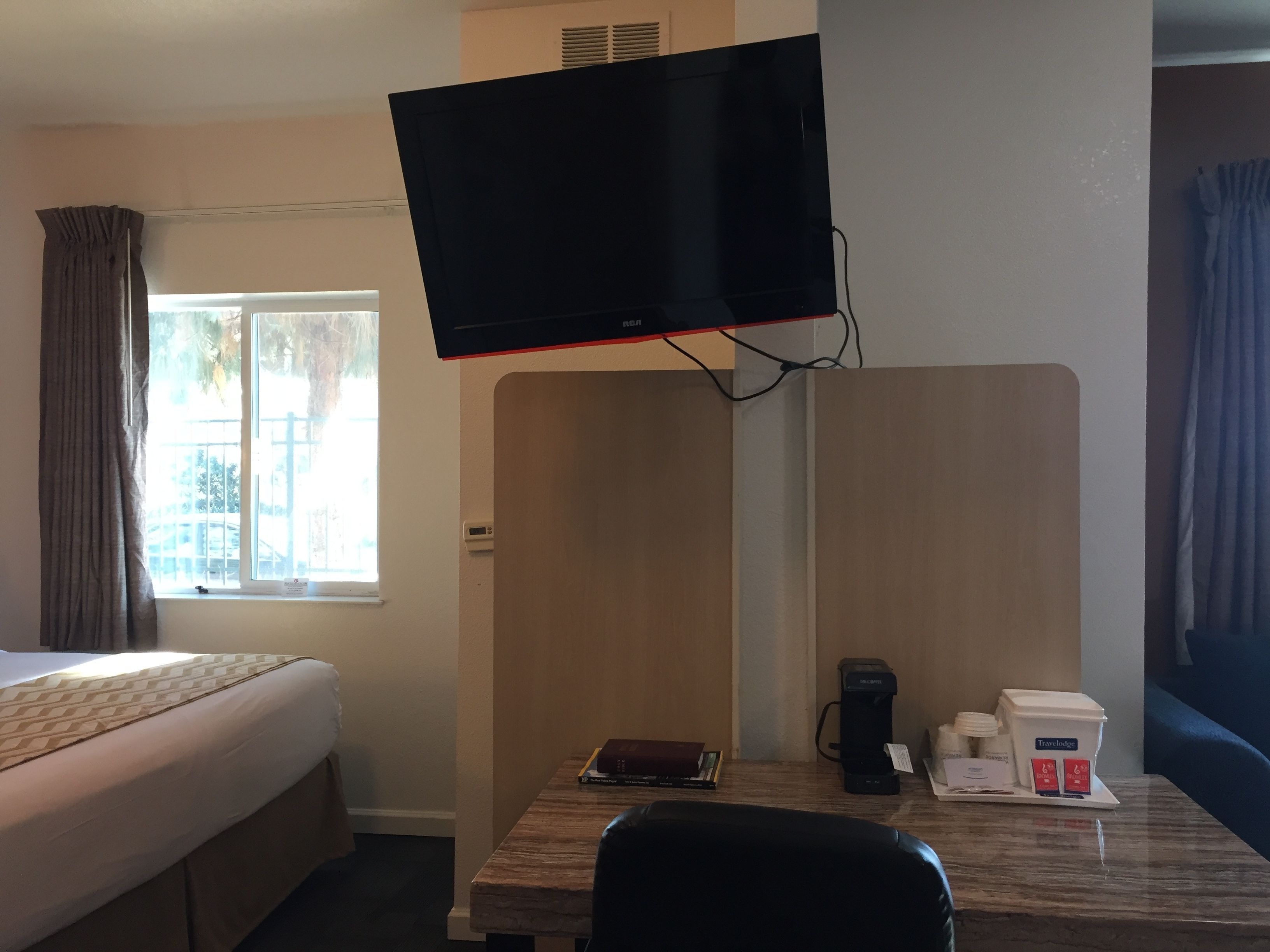 Quarto de hotel com cama, escrivaninha com cafeteira, TV na parede e janela com cortinas.