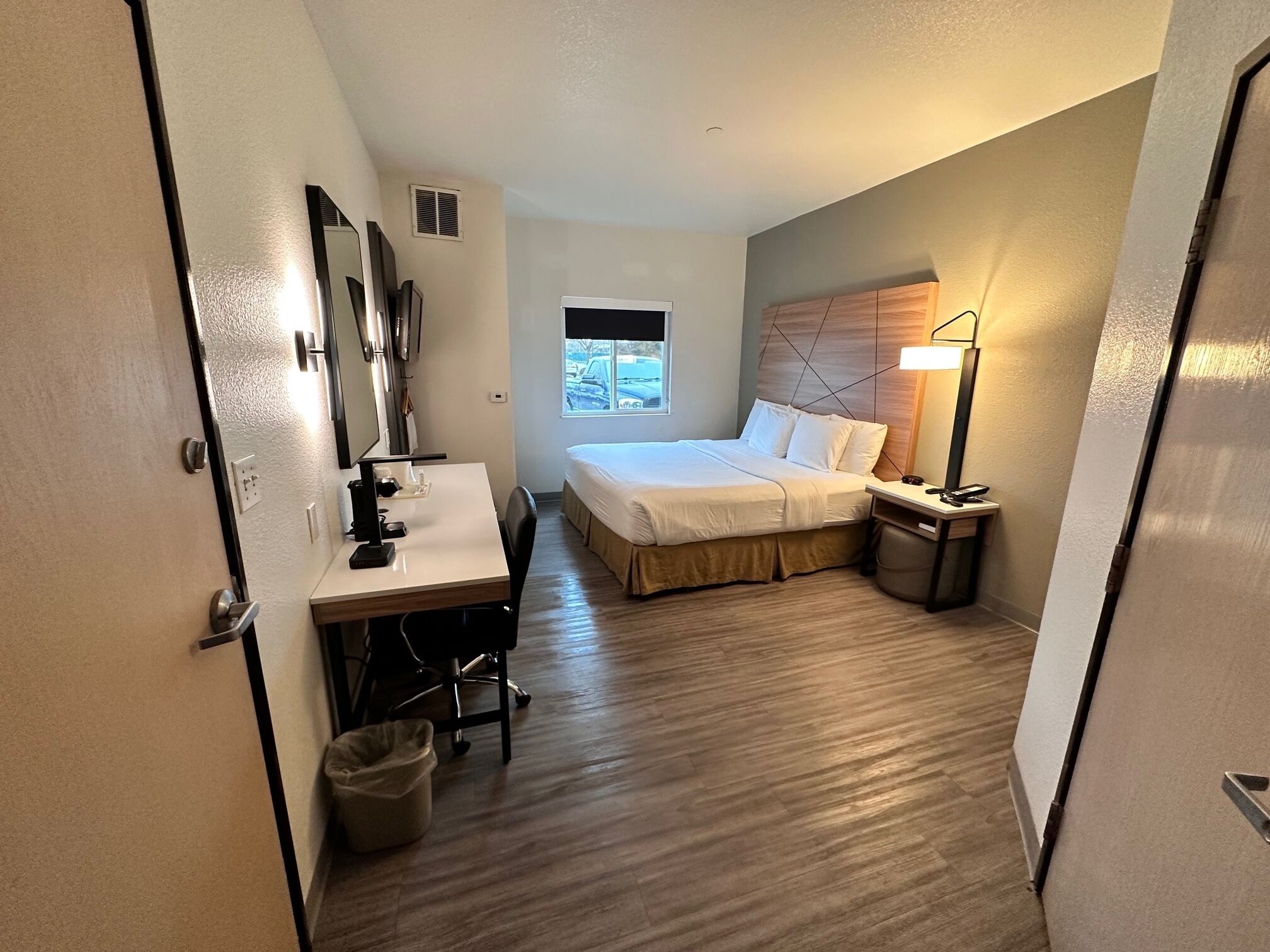 Una habitación de hotel compacta con una cama, un escritorio con silla, una lámpara de escritorio, un cesto de basura y una ventana.