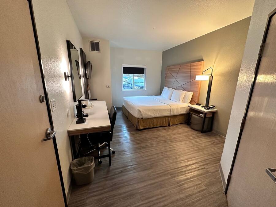 A compact hotel room with a bed, a desk with a chair, a desk lamp, a trash bin, and a window.