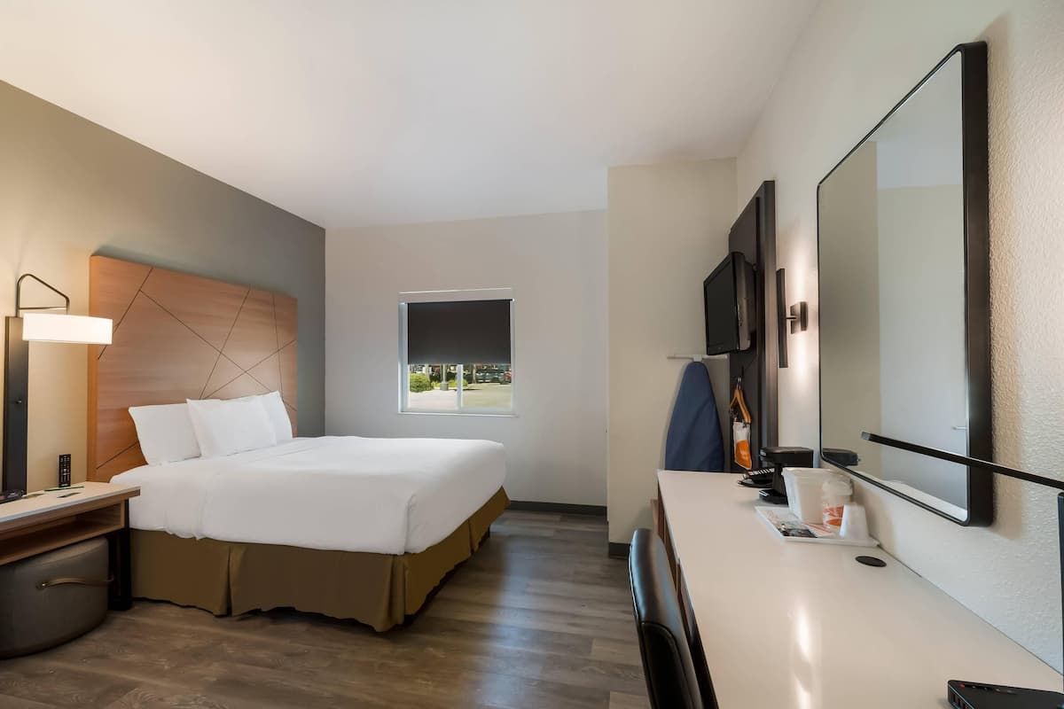 Quarto de hotel com cama, mesa de cabeceira, luminária, televisão e um espelho grande.