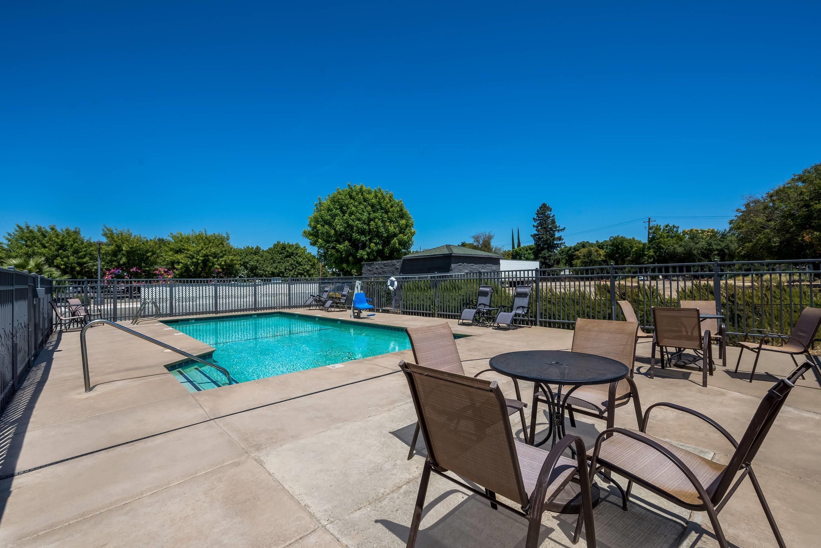 A swimming pool with lounge chairs and a table, surrounded by a fence and greenery.