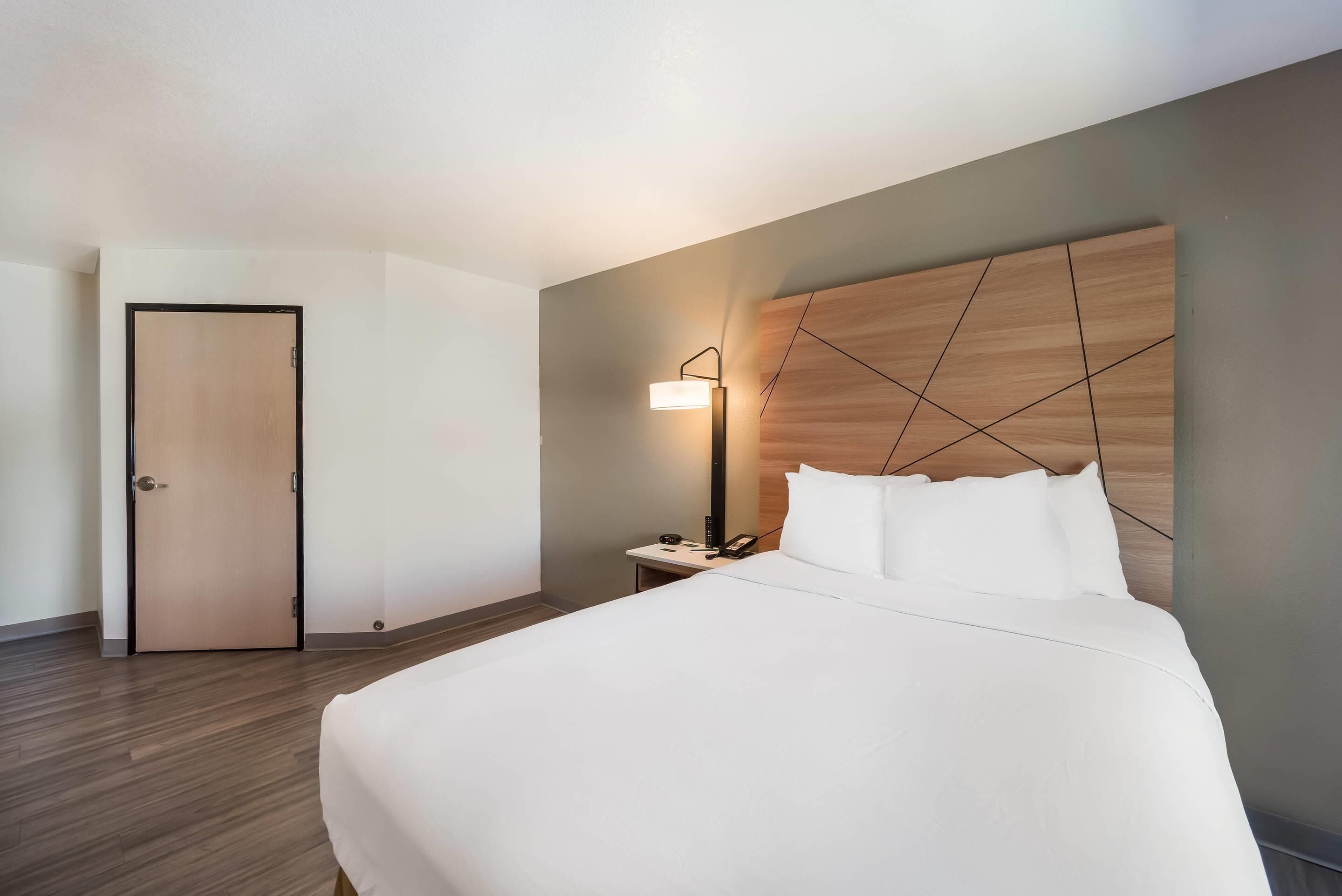 A hotel room with a bed, a bedside table with a lamp, a wooden headboard, and a door.
