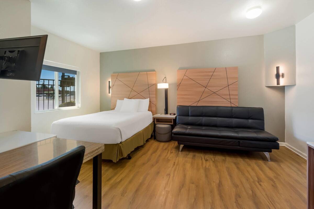 Quarto de hotel com cama, escrivaninha, sofá de couro preto, cabeceira de madeira, televisão fixada na parede e janela com vista para um estacionamento.