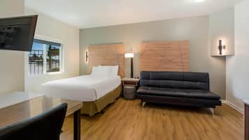 A hotel room with a bed, a desk, a black leather sofa, a wooden headboard, a wall-mounted TV, and a window with a view of a parking lot.