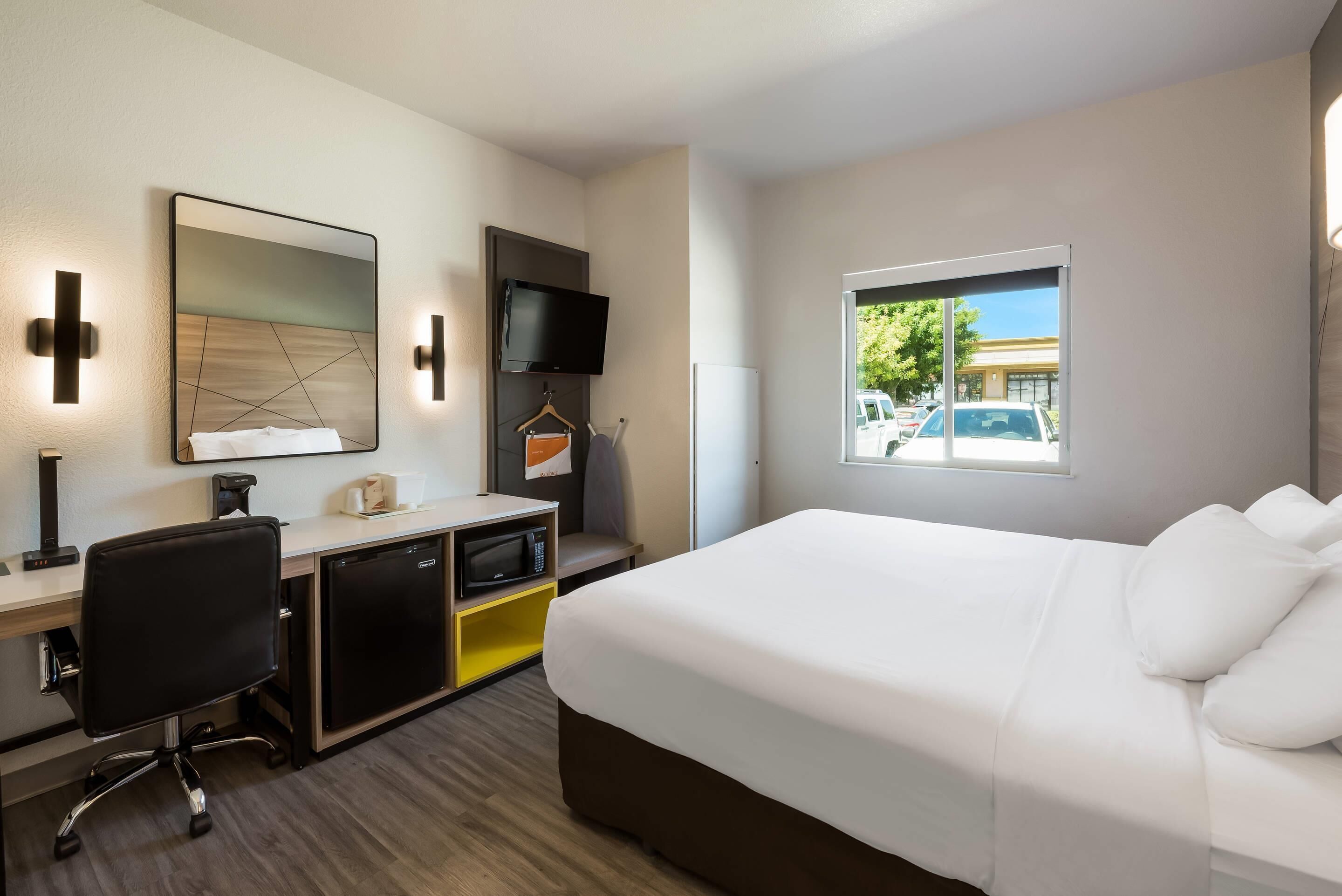 A hotel room with a bed, desk, chair, TV, and a window with a view of parked cars.