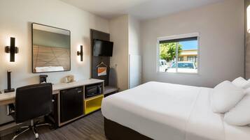 A hotel room with a bed, desk, chair, TV, and a window with a view of parked cars.