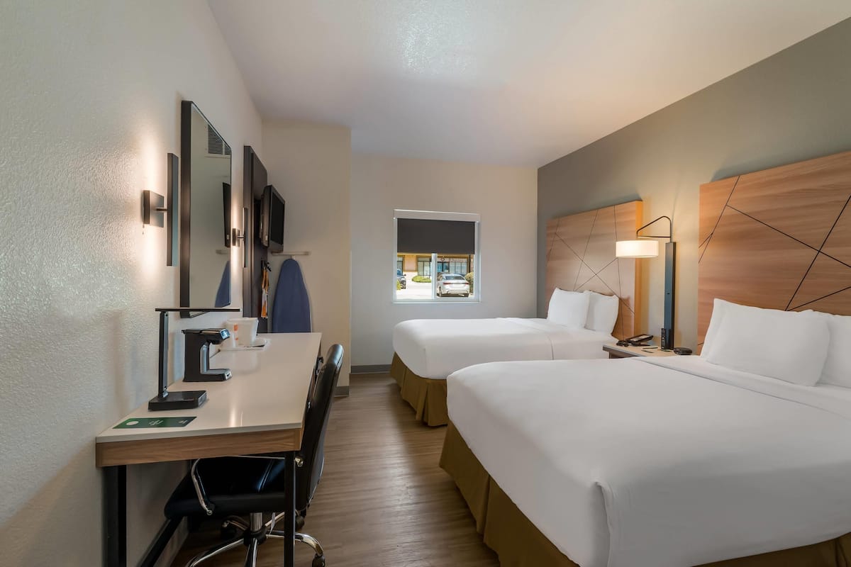 Quarto de hotel com duas camas, uma escrivaninha com cadeira, um espelho, um abajur e uma janela com vista para o estacionamento.