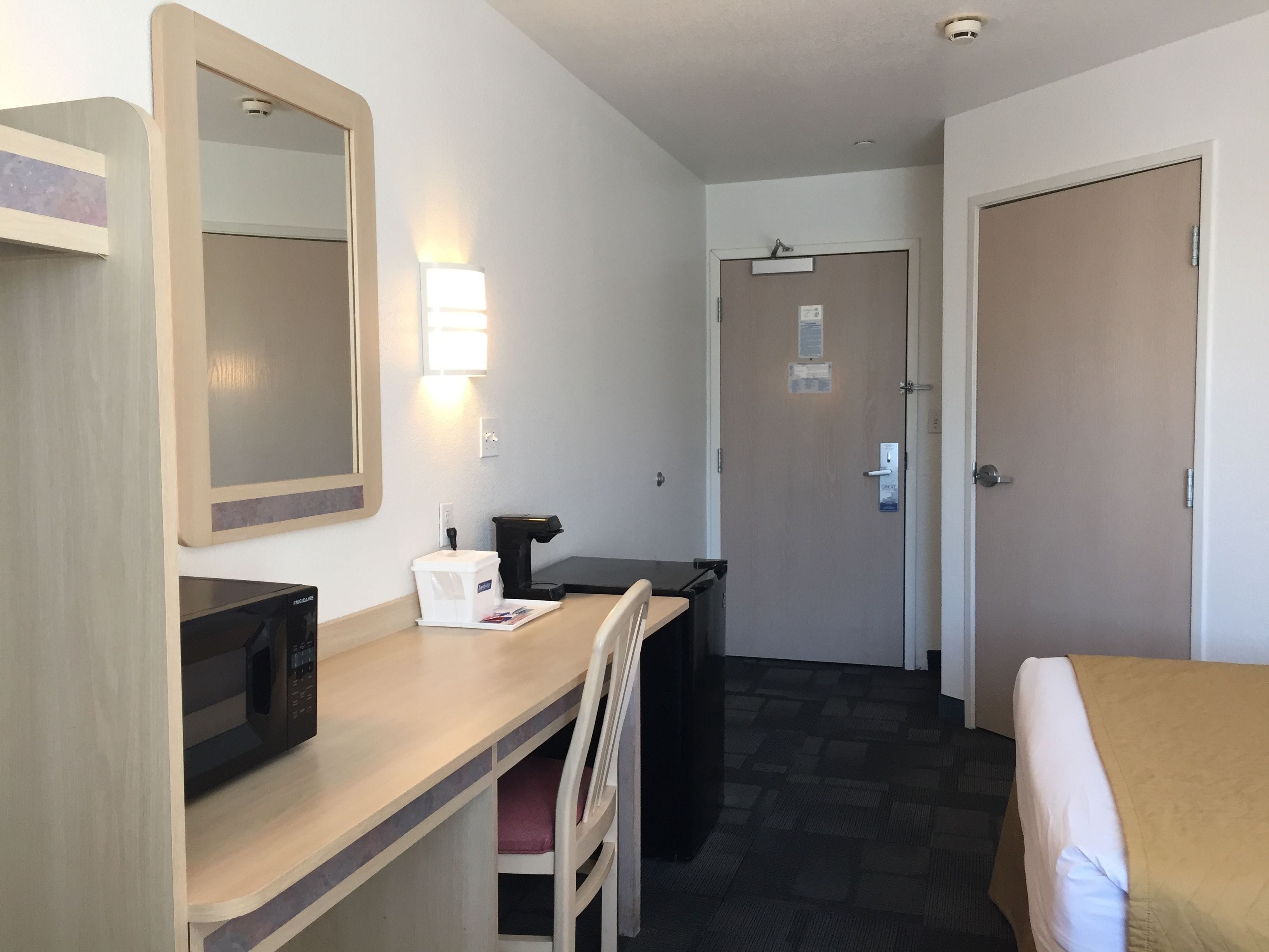 A hotel room with a bed, a desk with a microwave, a chair, a trash bin, and a door.