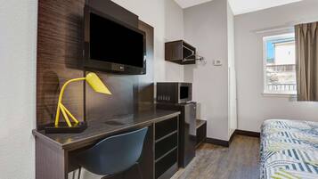 Habitación de hotel con escritorio de madera, una silla azul, cama con ropa de cama estampada, televisor, microondas y ventana con cortinas.