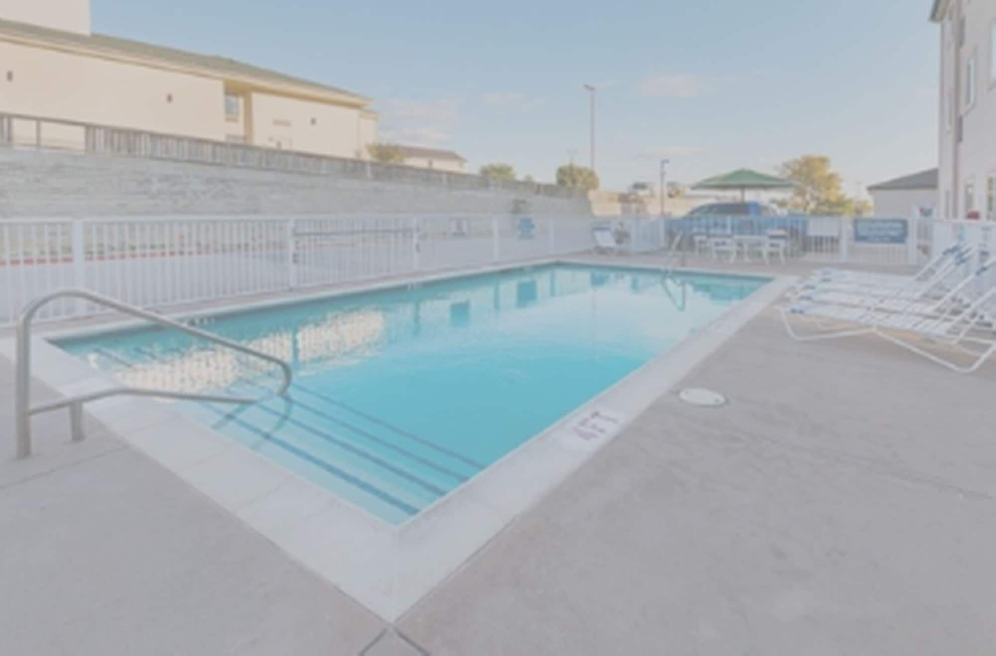 Una piscina rectangular con una escalera metálica en un lado y una zona de terraza con baldosas y sillas de descanso.