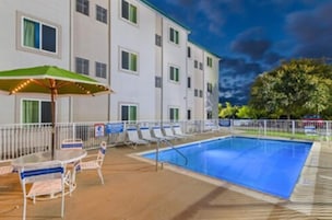 Zona de piscina de hotel con una piscina rectangular, sillas de descanso y una mesa con sombrilla verde.