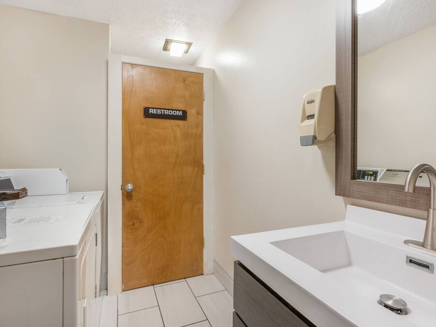 A bathroom with a wooden door labeled 'RESTROOM,' a white sink with a modern faucet, and a hand soap dispenser on the wall.
