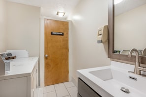 Un baño con una puerta de madera con la leyenda 'RESTROOM', un lavamanos blanco con un grifo moderno y un dispensador de jabón en la pared.