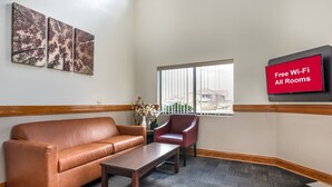 A hotel lobby with a brown leather sofa, a wooden coffee table, a red armchair, and a wall-mounted TV displaying "Free Wi-Fi All Rooms". - Red Roof Inn Greencastle South – Cloverdale (Cloverdale)