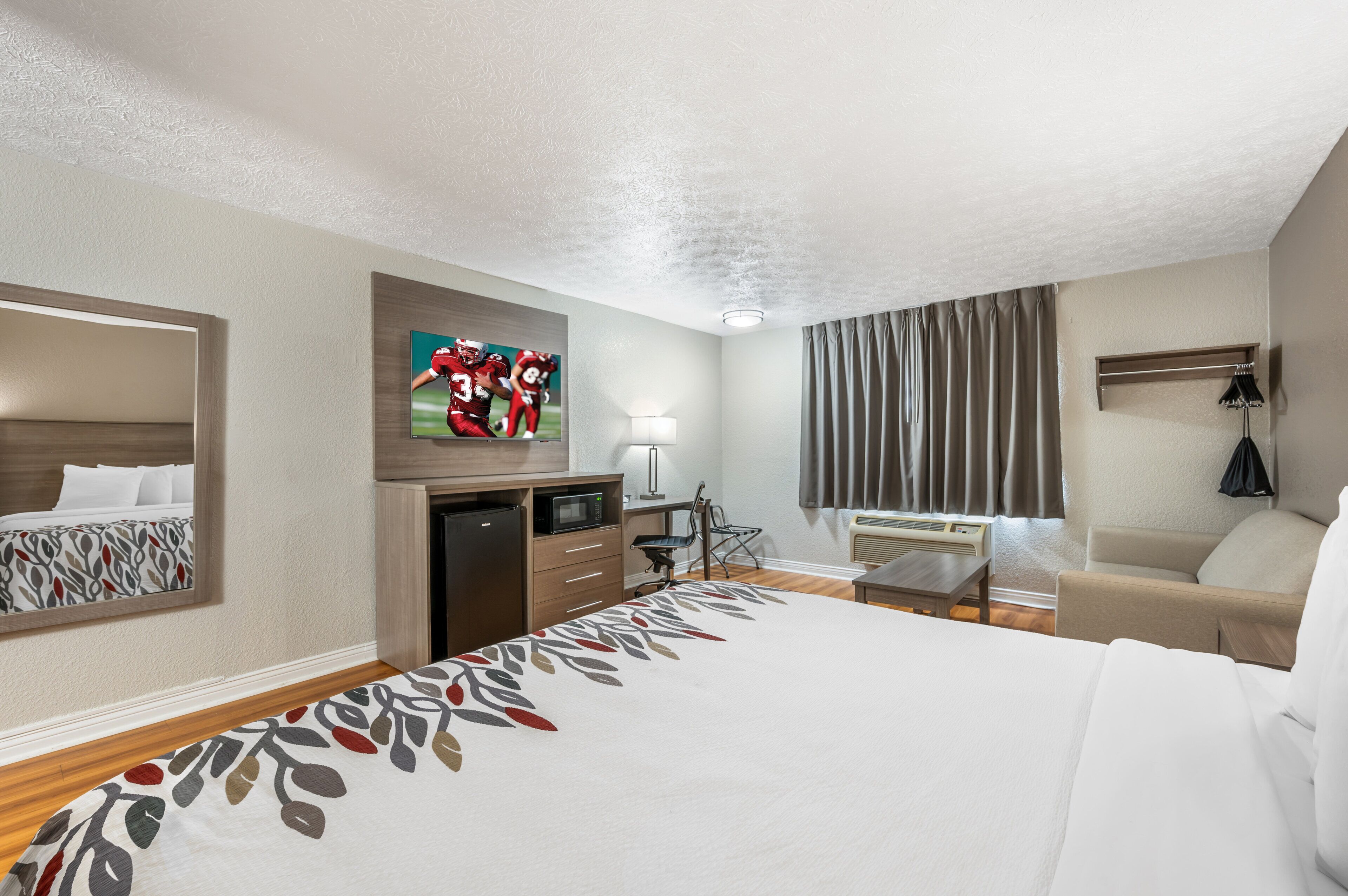 A hotel room with a bed, a desk, a chair, a microwave, a refrigerator, a television, a window with curtains, and a hanging bag.