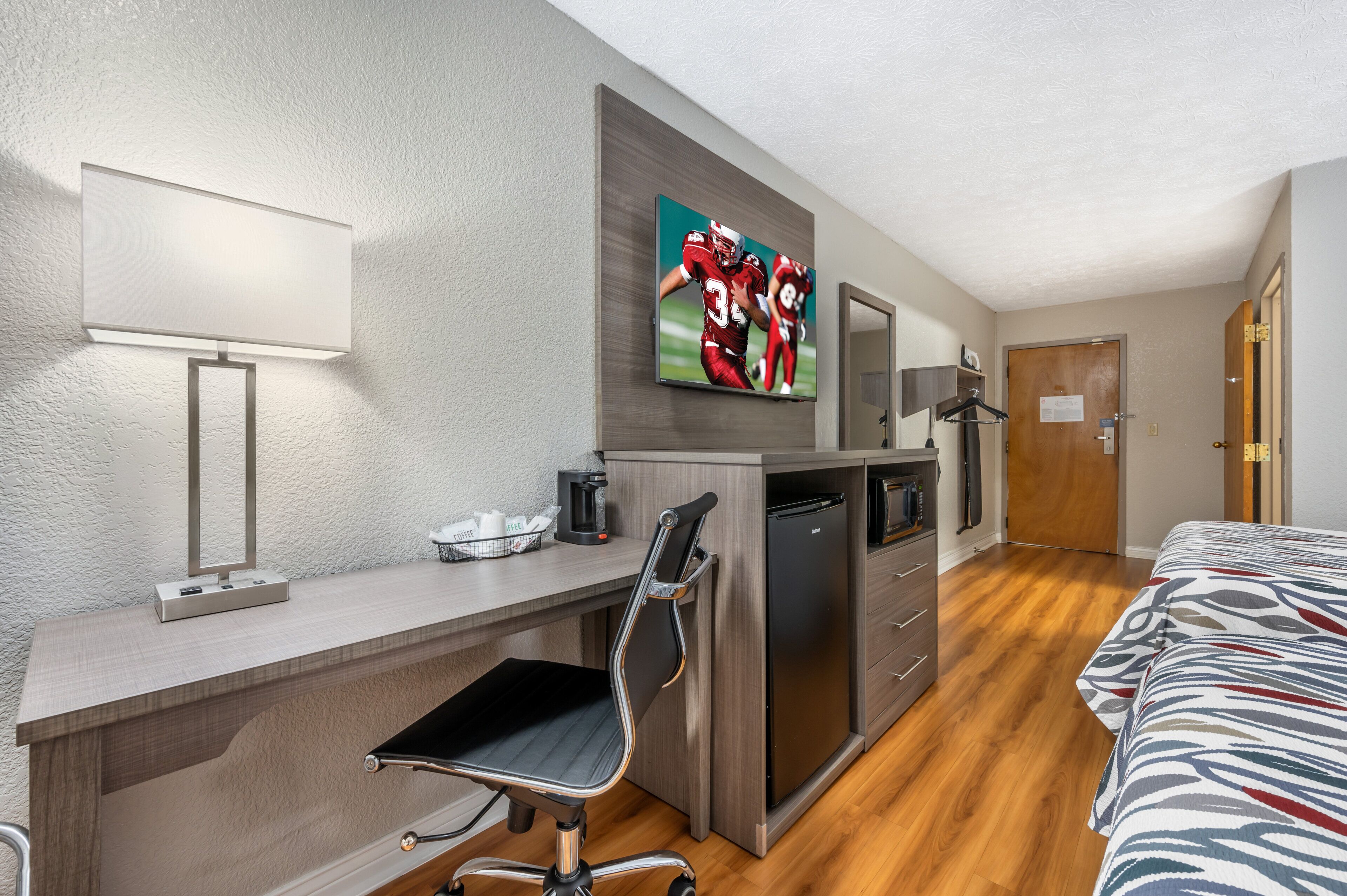 Ein Hotelzimmer mit Schreibtisch, einer Kaffeemaschine, einem Kühlschrank, einem Bett und einem Fernseher, der ein Sportspiel zeigt.