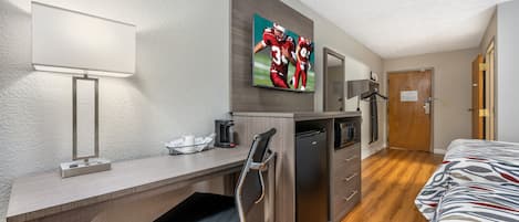 Habitación de hotel con escritorio, cafetera, refrigerador, cama y televisor mostrando un partido deportivo.