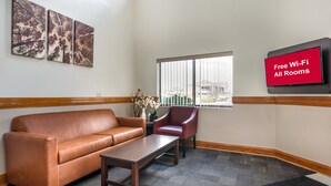 A hotel lobby with a leather sofa, a wooden coffee table, a red armchair, a vase with flowers, and a wall-mounted TV displaying 'Free Wi-Fi All Rooms'. - Red Roof Inn Greencastle South – Cloverdale (Cloverdale)