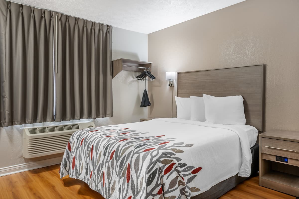 Una habitación de hotel con cama, mesita de noche, lámpara, aire acondicionado y cortinas.