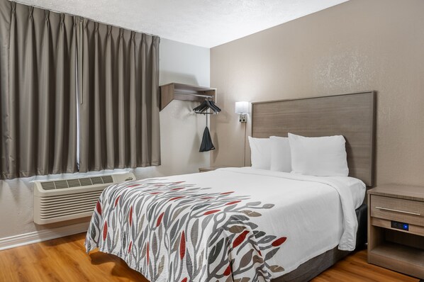 A hotel room with a bed, a nightstand, a lamp, a window with curtains, and an air conditioning unit. - Red Roof Inn Greencastle South – Cloverdale (Cloverdale)
