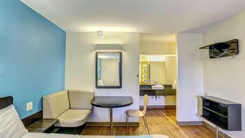 A hotel room with a bed, a small desk, a chair, a mirror, a TV, and a bathroom visible in the background.