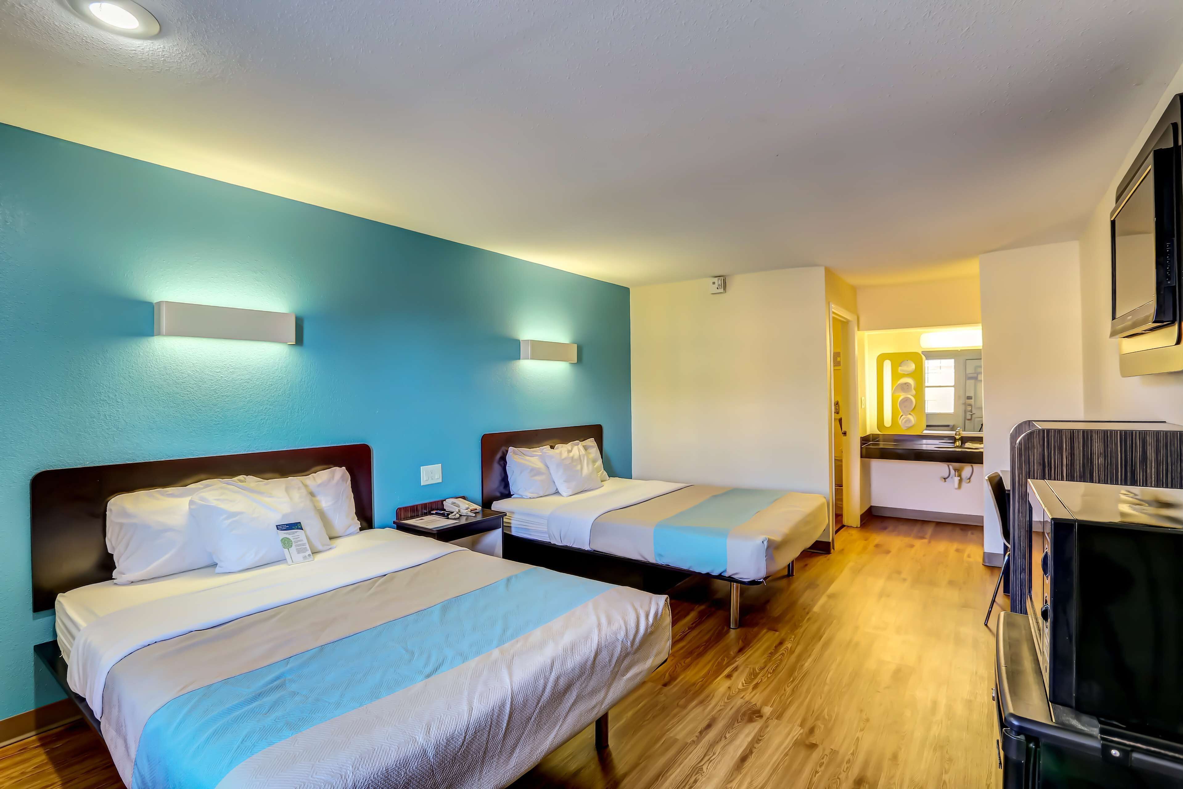 A hotel room with two beds, a wooden floor, a TV, and a kitchenette in the background.