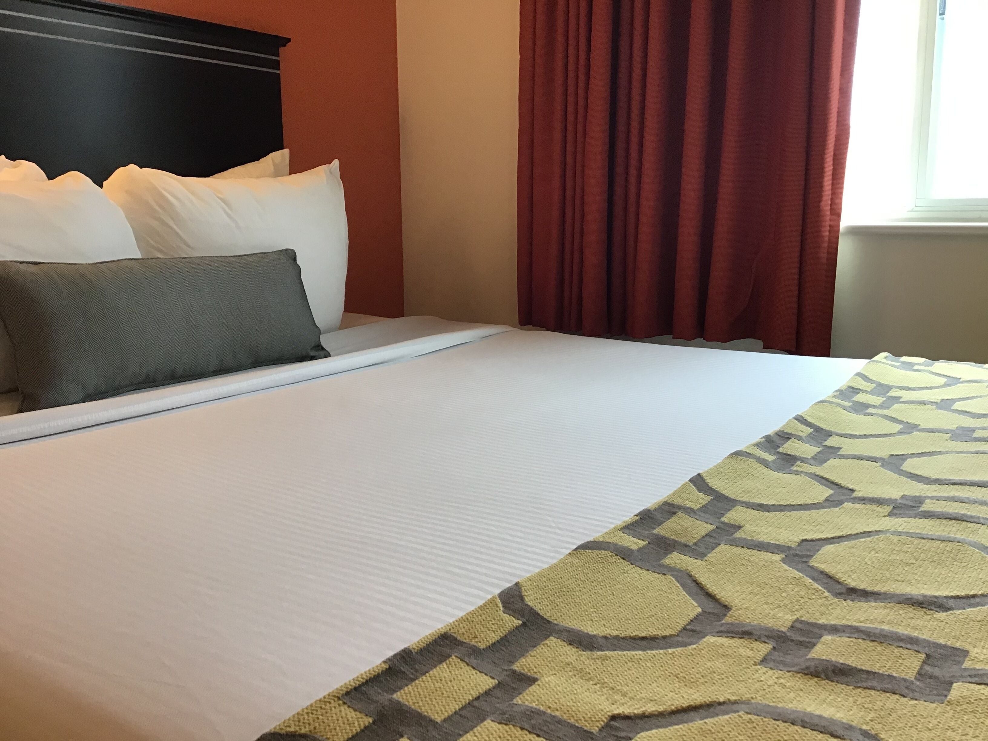 A hotel room with a neatly made bed, a patterned throw blanket, and red curtains.