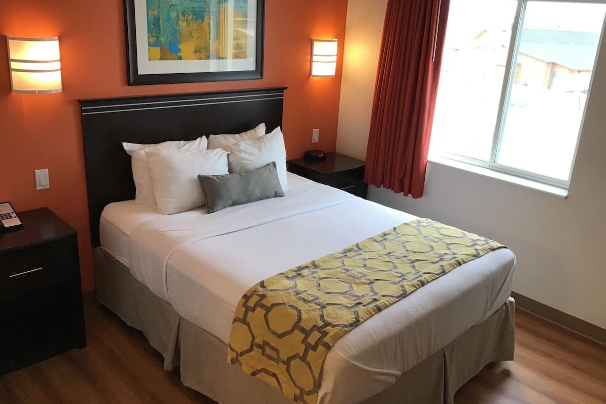 A neatly made bed with a patterned bedspread, two bedside lamps, a framed artwork above the bed, and a window with curtains.