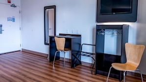 A modern room with a wooden floor, a desk with two chairs, a flat-screen TV mounted on the wall, and a coat rack with hanging items. - Motel 6 Pottstown, PA (Pottstown)