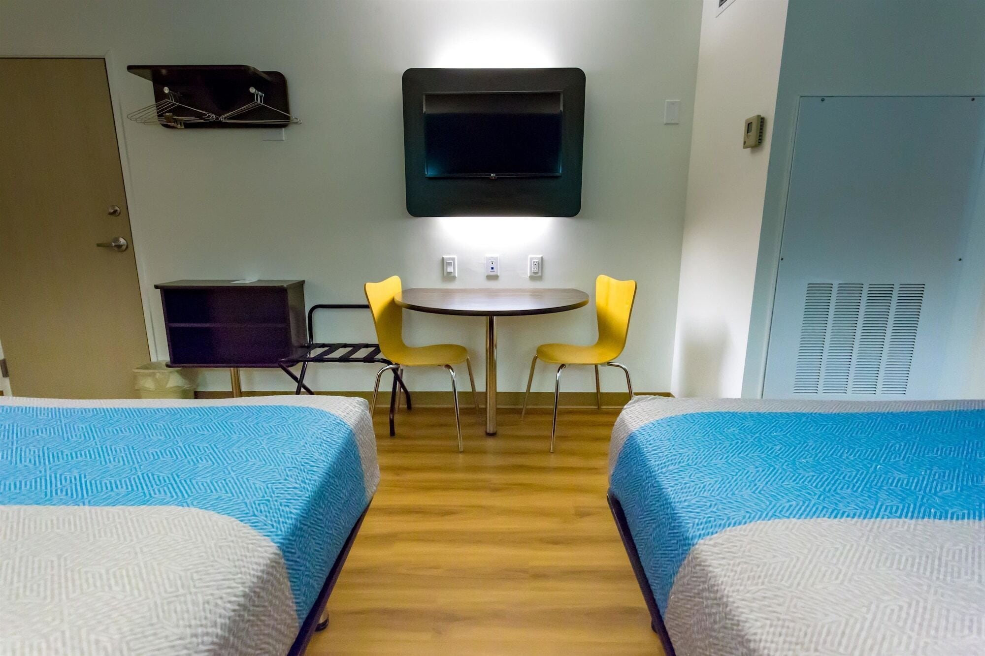 A hotel room with two beds, a small table with two chairs, a TV mounted on the wall, and a wardrobe.