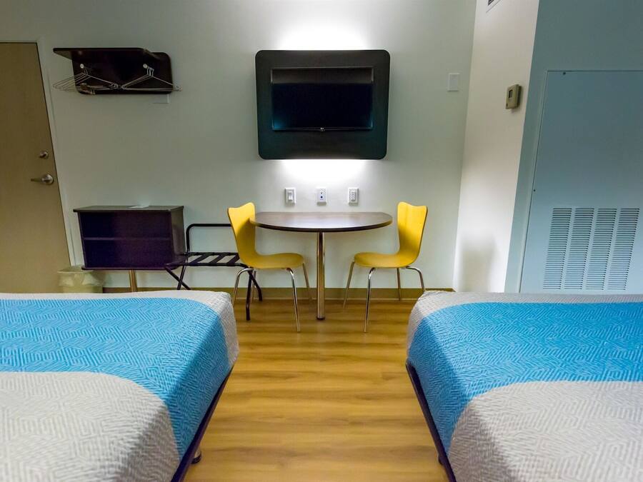 A hotel room with two beds, a small table with two chairs, a TV mounted on the wall, and a wardrobe.