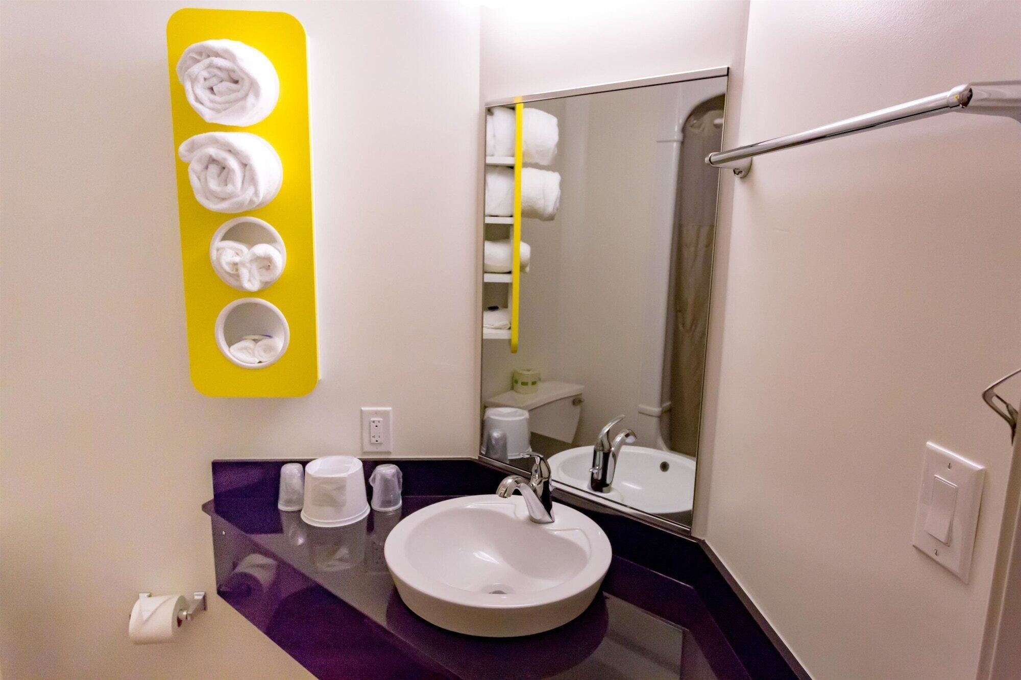 Un baño con un lavamanos blanco, un espejo, un perchero con tres toallas y una encimera morada.