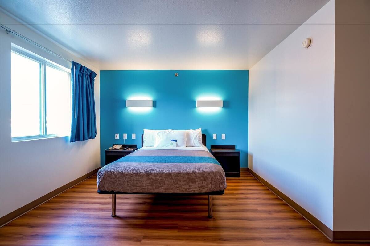 A hotel room with a bed, bedside tables, a nightstand, a wall-mounted light, and a window with curtains.