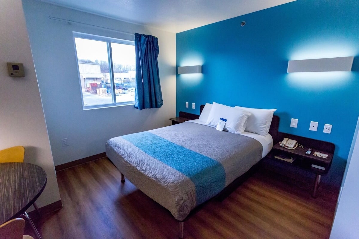 A hotel room with a bed, a nightstand, a chair, a window with curtains, and a view of the outside.