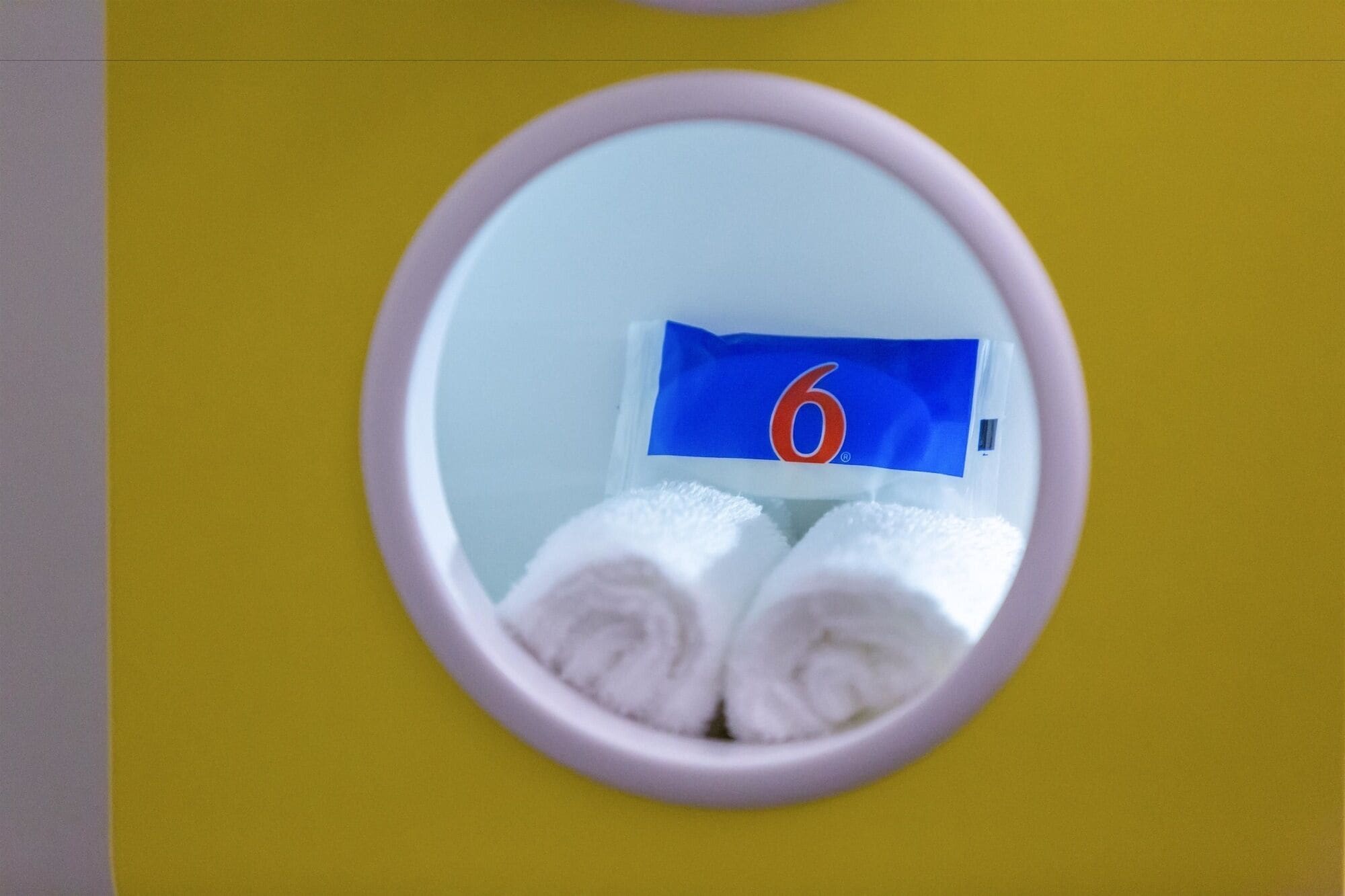 Espejo de baño con una toalla y un paquete azul etiquetado '6' sobre una pared amarilla.