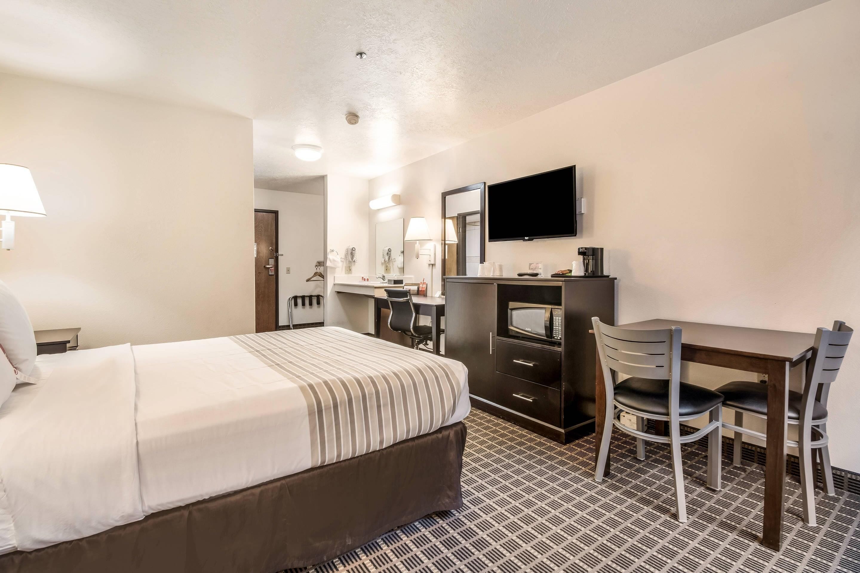 Quarto de hotel com cama, escrivaninha, duas cadeiras, televisão e micro-ondas.