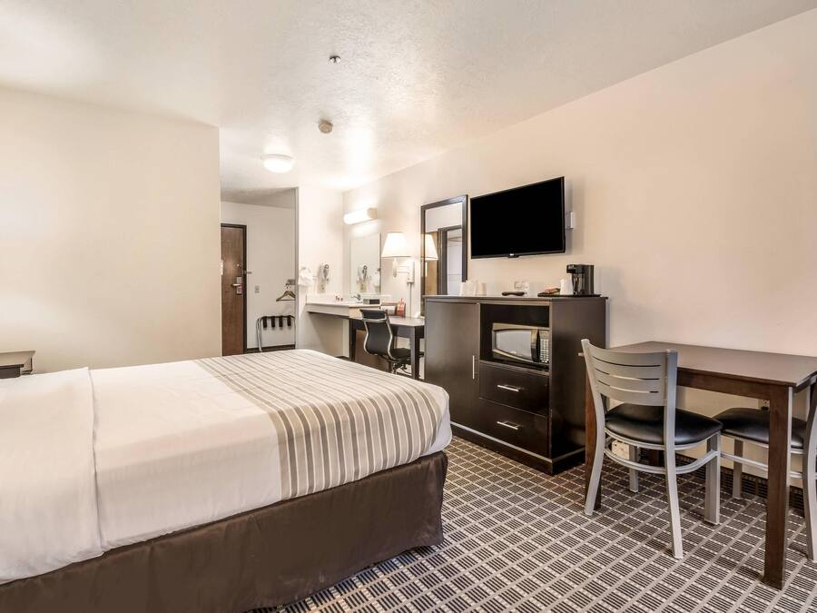 Quarto de hotel com cama, escrivaninha, duas cadeiras, televisão e micro-ondas.