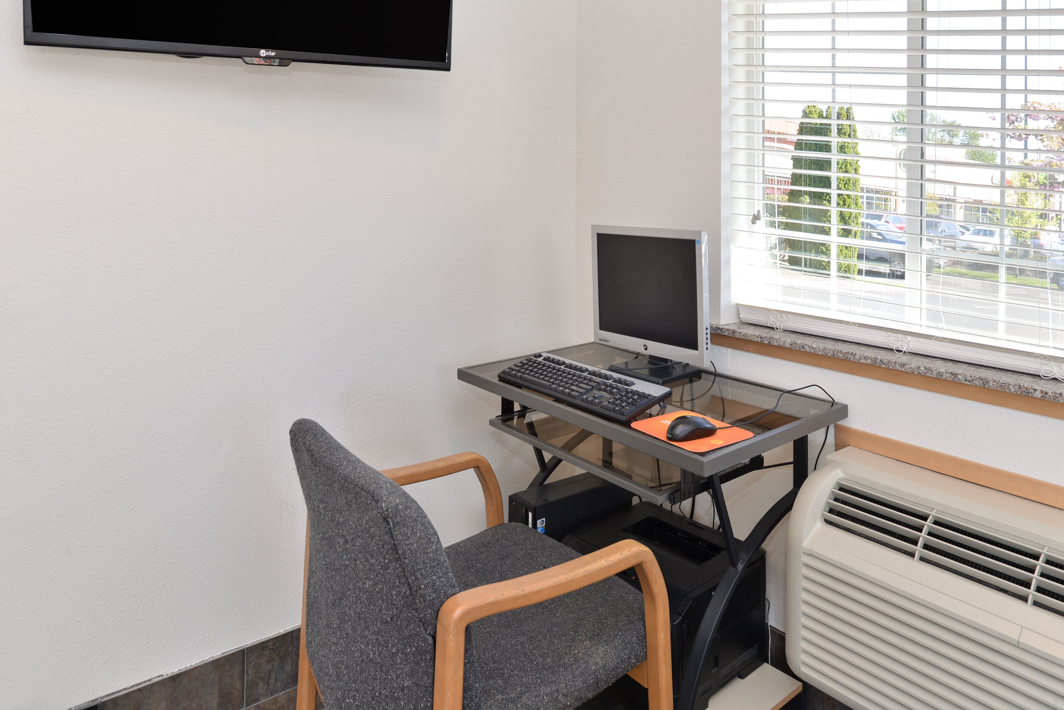 A computer desk with a monitor, keyboard, and mouse, a chair, a television mounted on the wall, and a window with blinds.