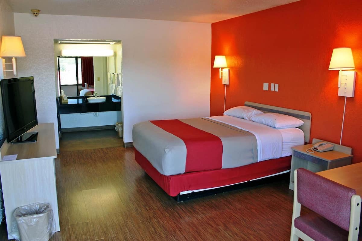 A hotel room with a bed, a desk with a television, a bathroom with a mirror, and two wall-mounted lamps.